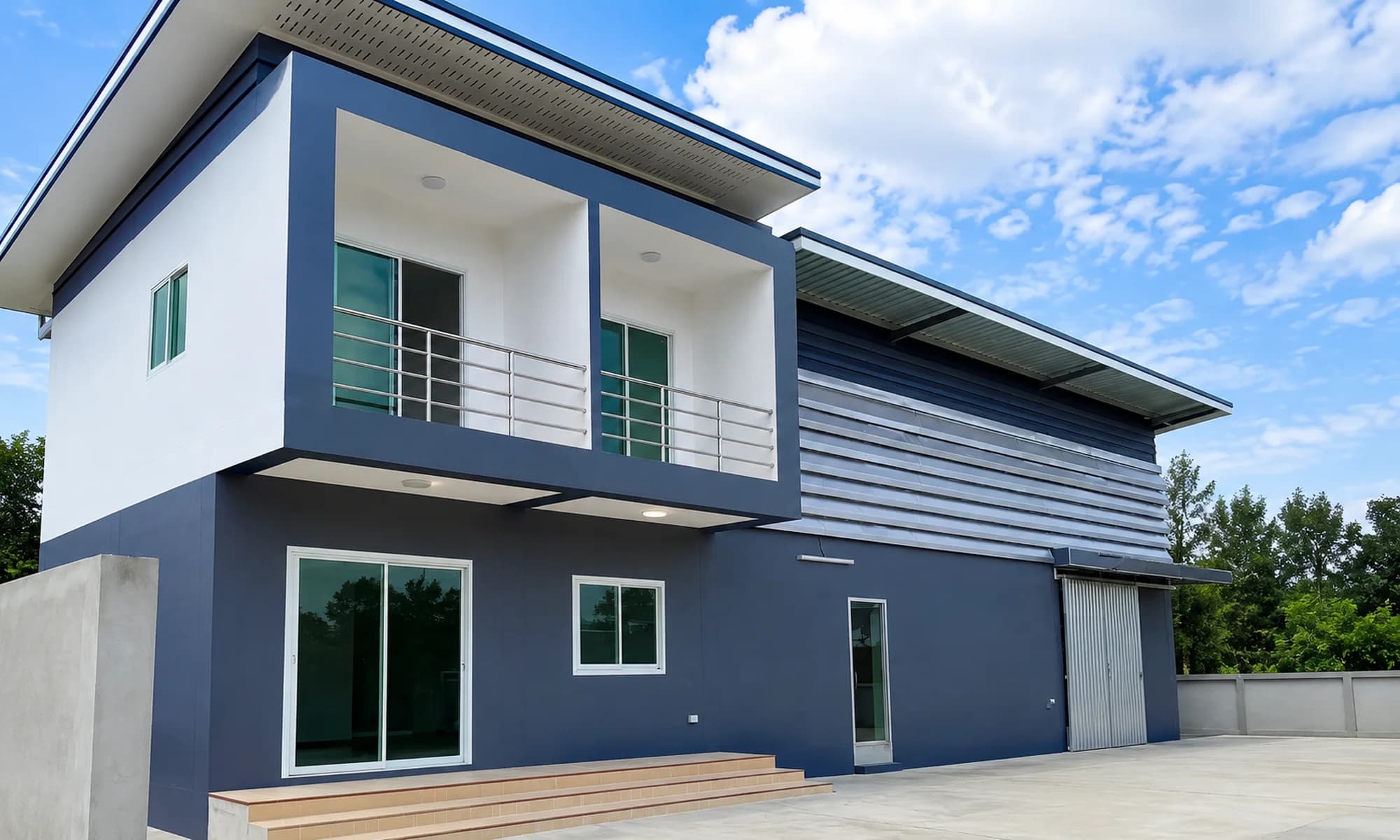 บ้านสองชั้นโทนน้ำเงิน–ขาวต่อเนื่องกับส่วนโรงงานด้านข้างบนลานคอนกรีต — โครงการ Wellgoodnet